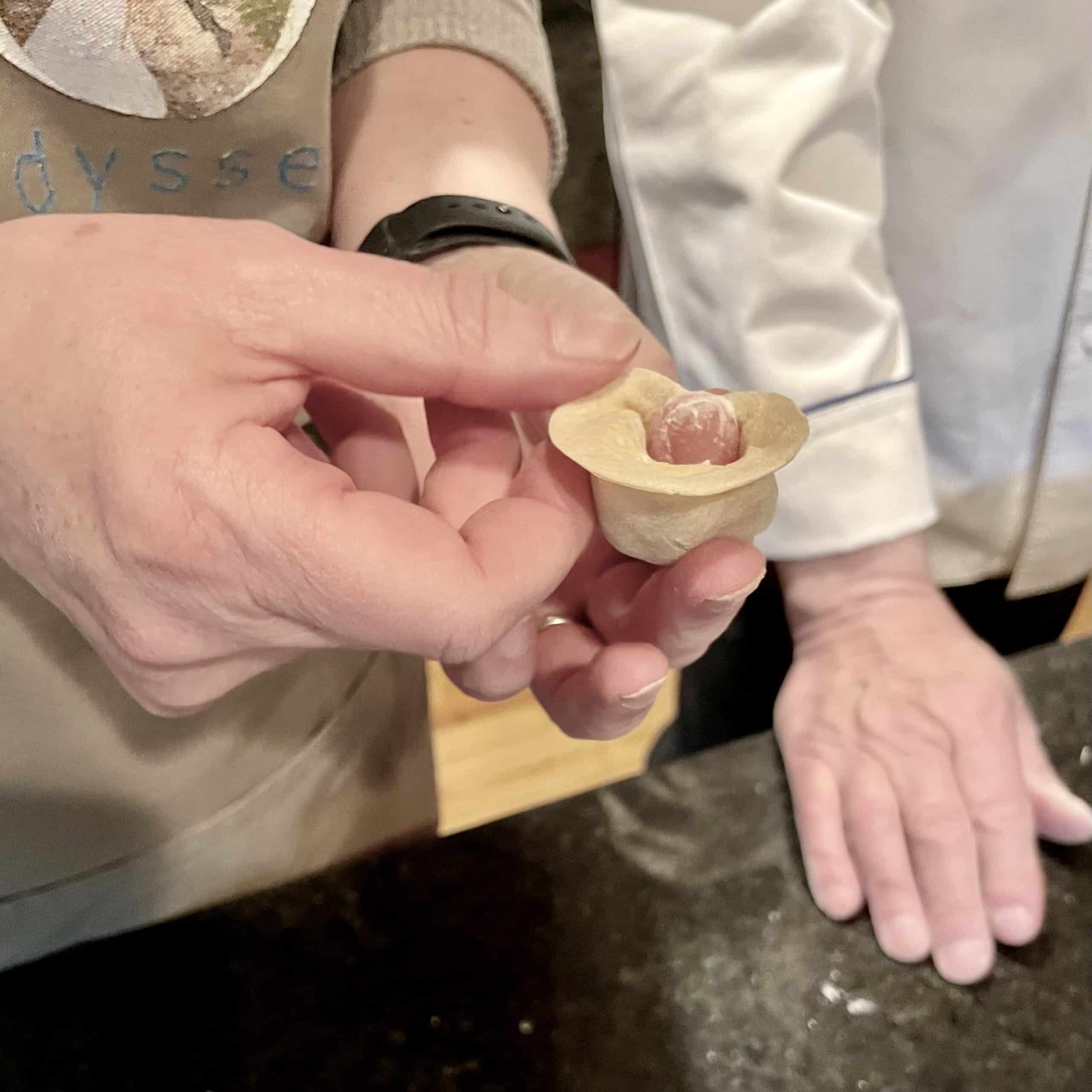Hand holding a small stuffed pasta piece, chef in background.