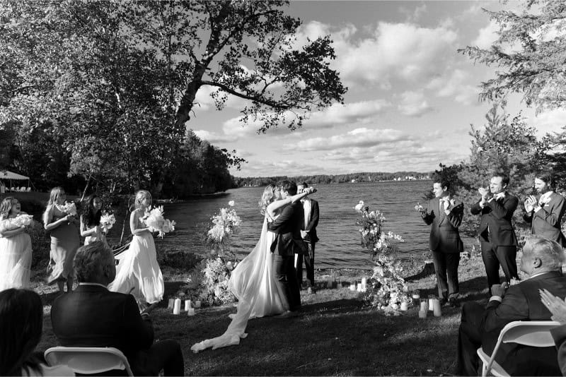 Outdoor wedding ceremony by the water with guests and floral decor.