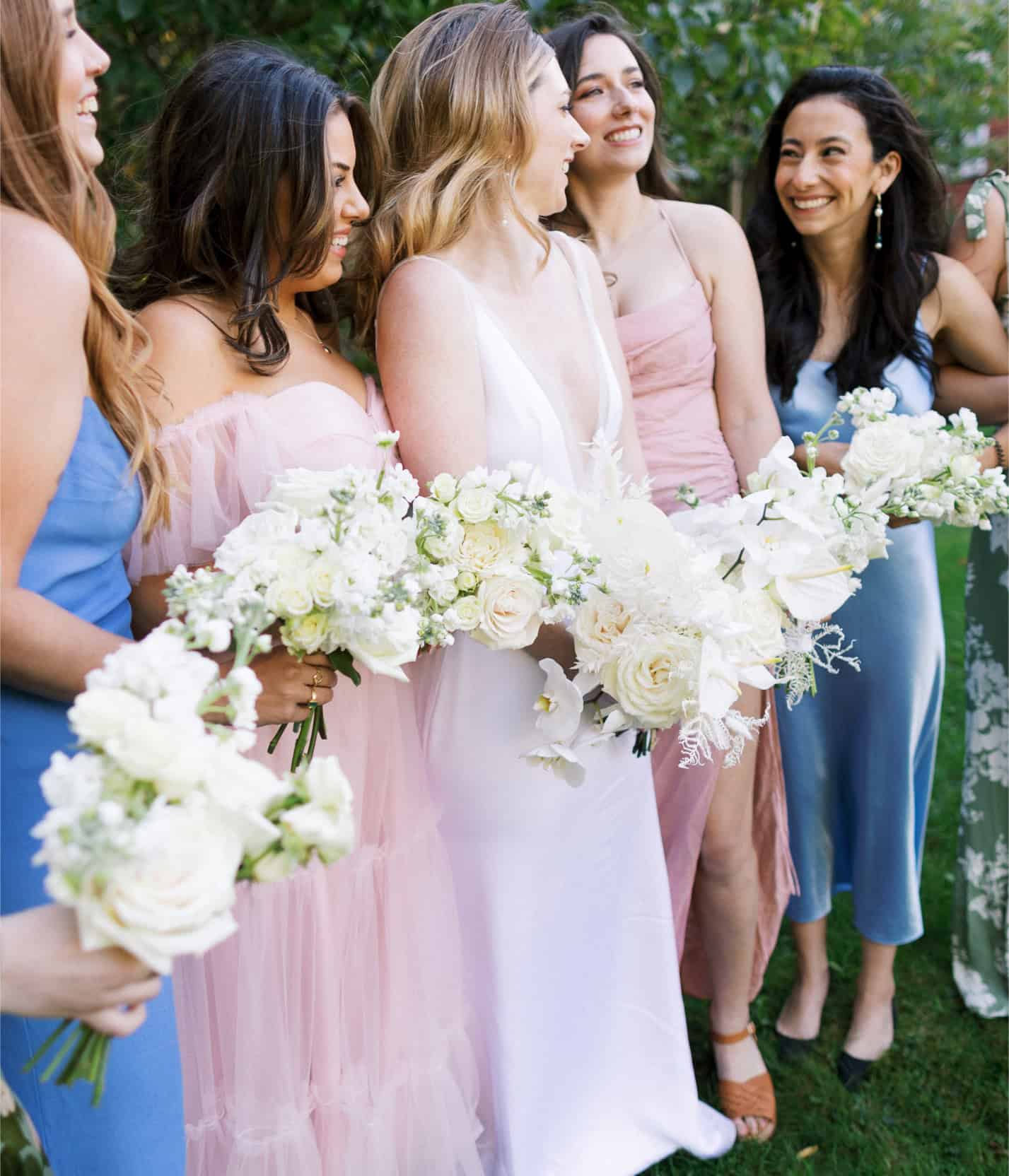 Bridal party with flowers, smiling women in pastel dresses at outdoor wedding.