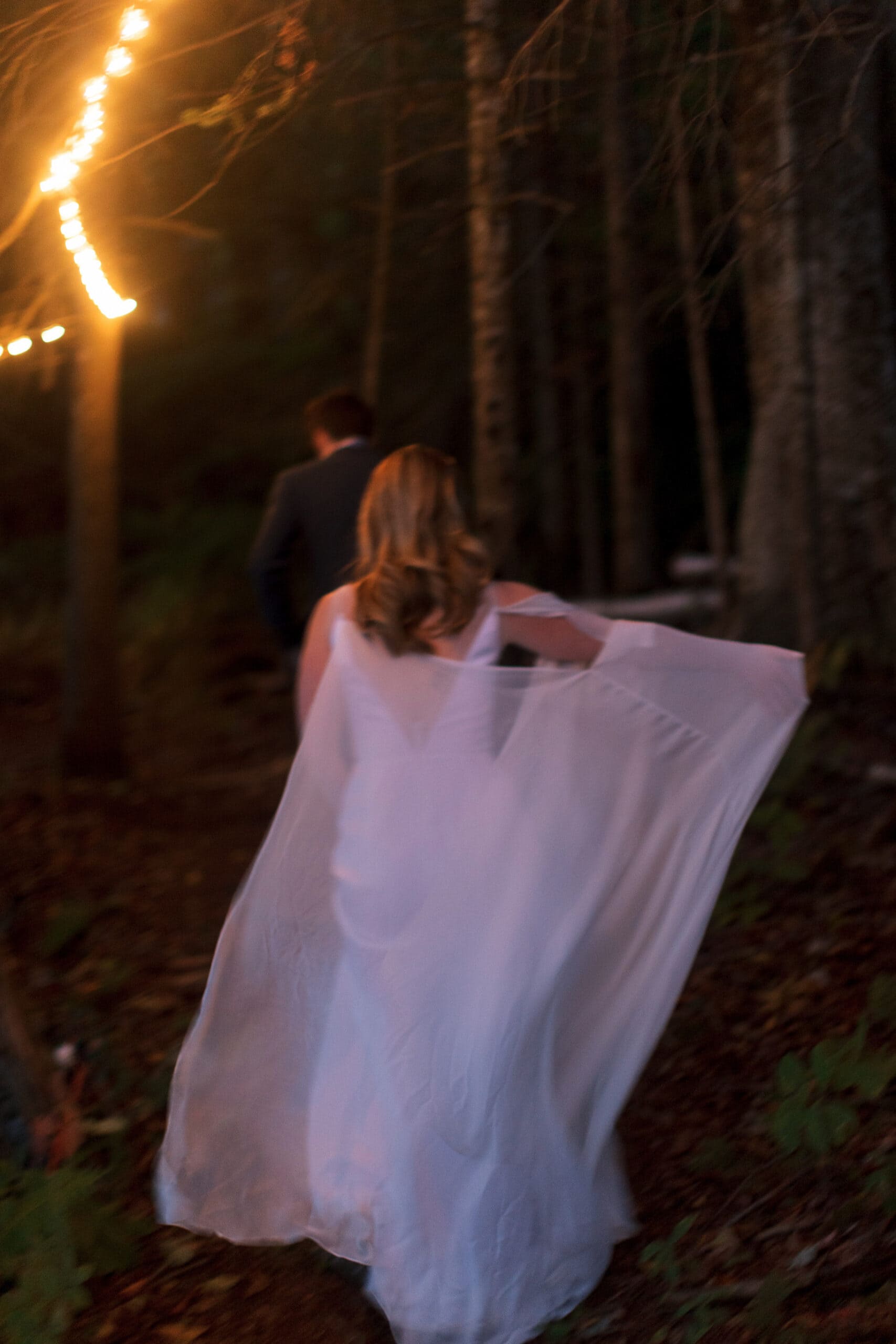 Bride walking through forest at dusk during outdoor wedding celebration.