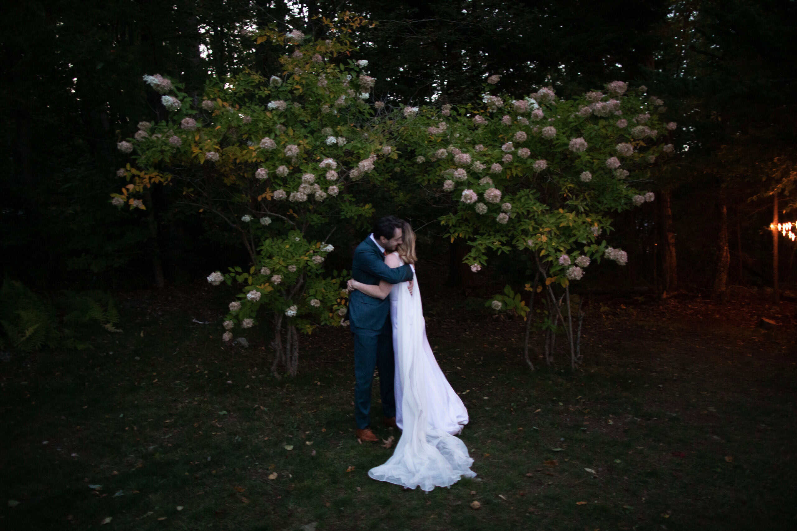 Outdoor wedding couple embracing beneath flowering trees at sunset.