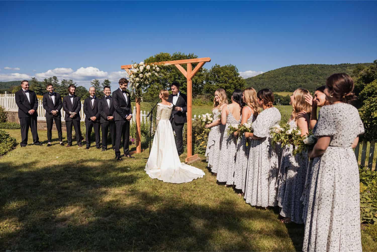 Outdoor wedding ceremony with bride, groom, bridesmaids, and groomsmen under a wooden arch.