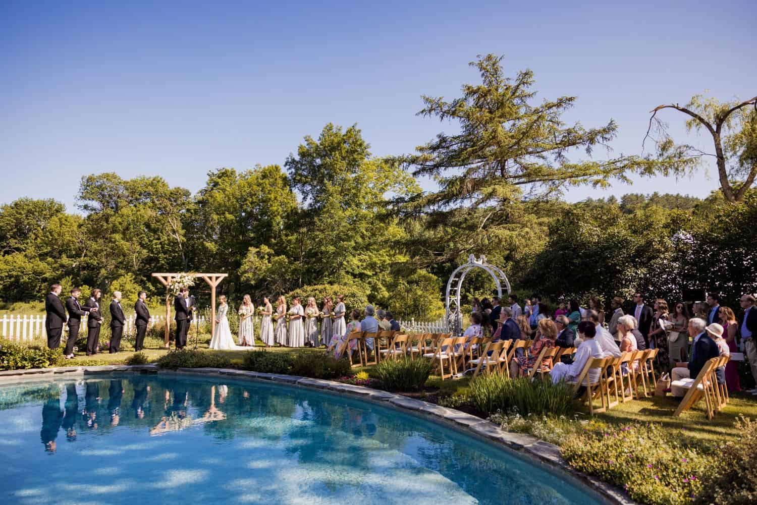 Outdoor wedding ceremony with guests, floral arch, and scenic garden backdrop.