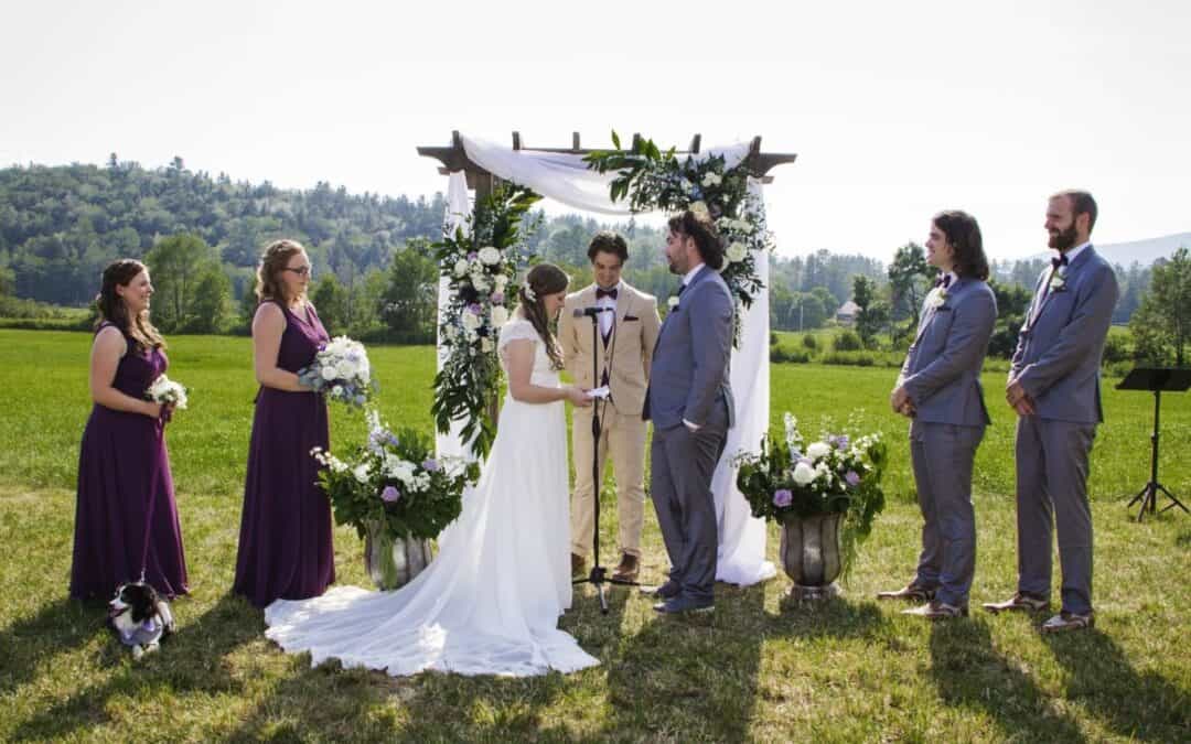 Outdoor wedding ceremony with bride and groom standing under floral arch, surrounded by bridesmaids.