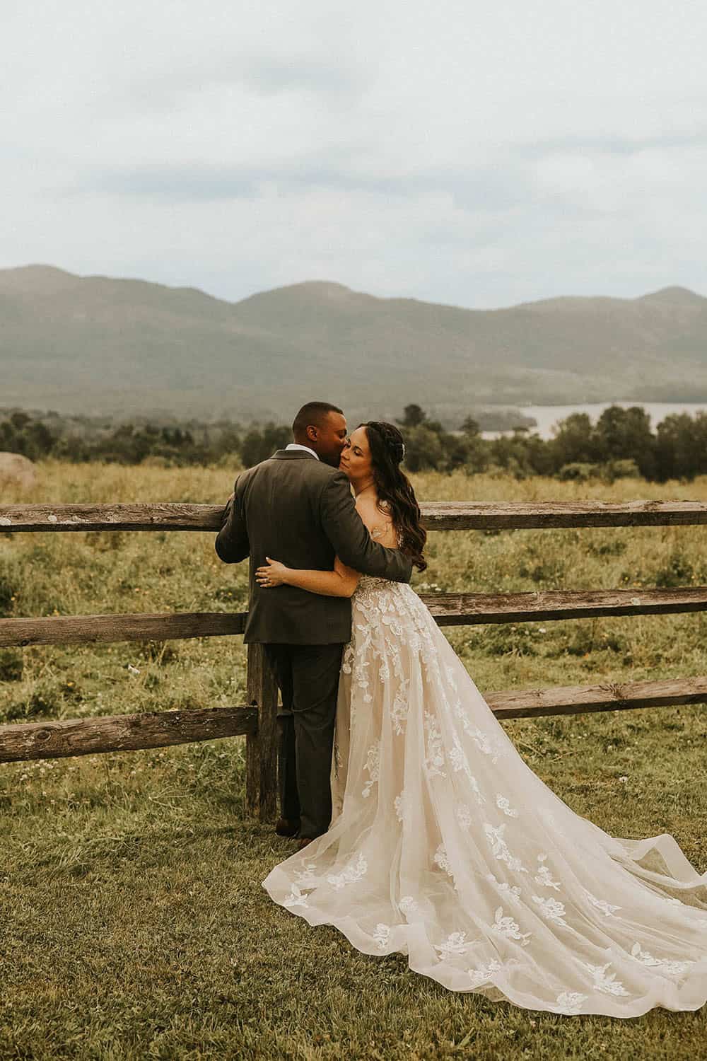 Romantic couple in wedding attire embracing outdoors with scenic mountain background.