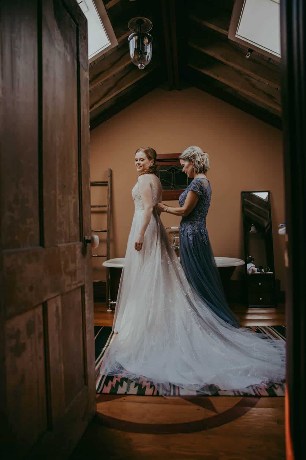 Bride in wedding gown with assistant helping her in a cozy, stylish room.