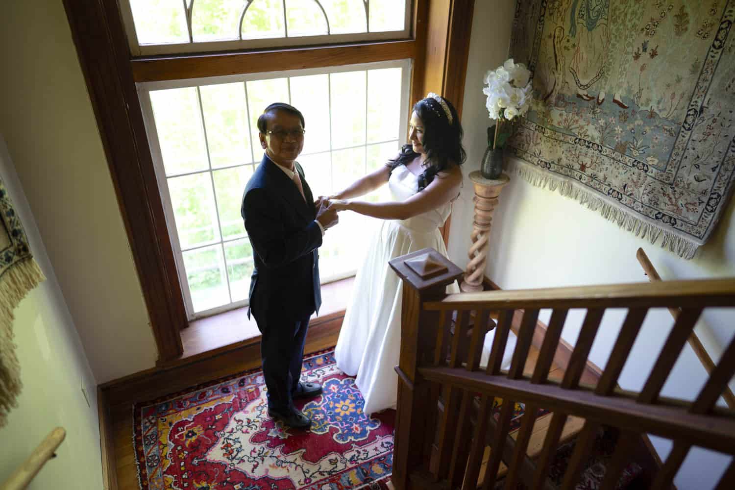 Bride and groom holding hands by a large window during their wedding.