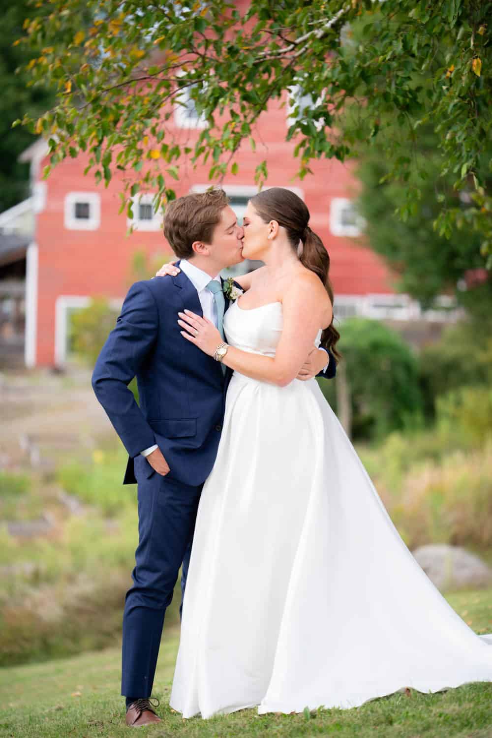 Wedding couple kissing in a scenic outdoor setting with greenery and a red barn in the background.