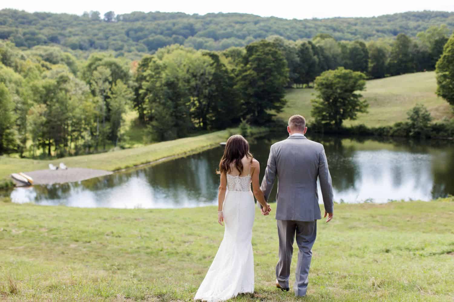 Wedding couple walking hand in hand near a peaceful pond in a lush green landscape.